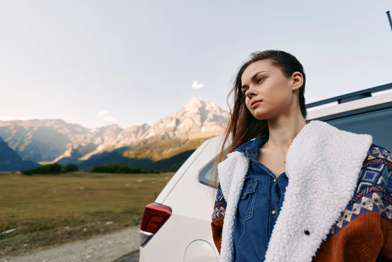 Woman by car in mountain landscape, travel portrait of a young woman leaning on a vehicle beside a rural road, scenic outdoor adventure with peaks, meadow and thoughtful expression
