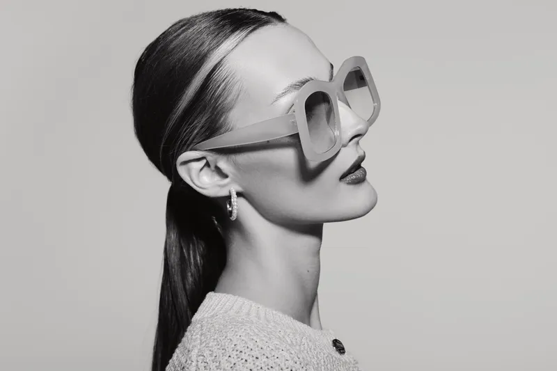 Stylish white woman with pale skin and slicked back hair wears oversized sunglasses and earrings, posing in profile on a neutral background. Concept for fashion, elegance, and modern style.