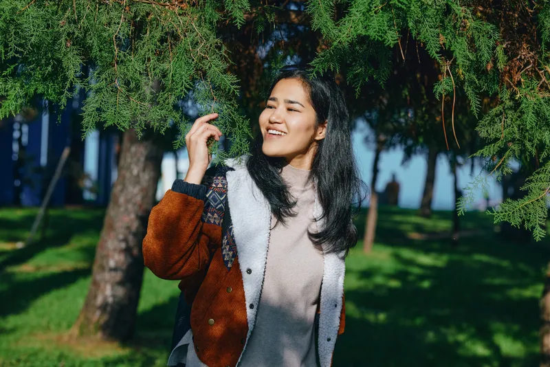 Woman with a warm smile in a sunny outdoor park scene, wearing a brown and white jacket and sweater, capturing relaxed mood, natural light, and casual fashion for lifestyle use today