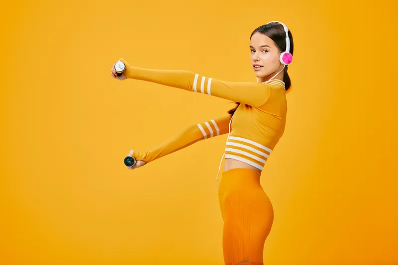 Woman in orange athletic wear stretches and lifts light dumbbells against a vivid orange backdrop, showcasing dynamic fitness, balance, and energy in a bold, studio workout scene.
