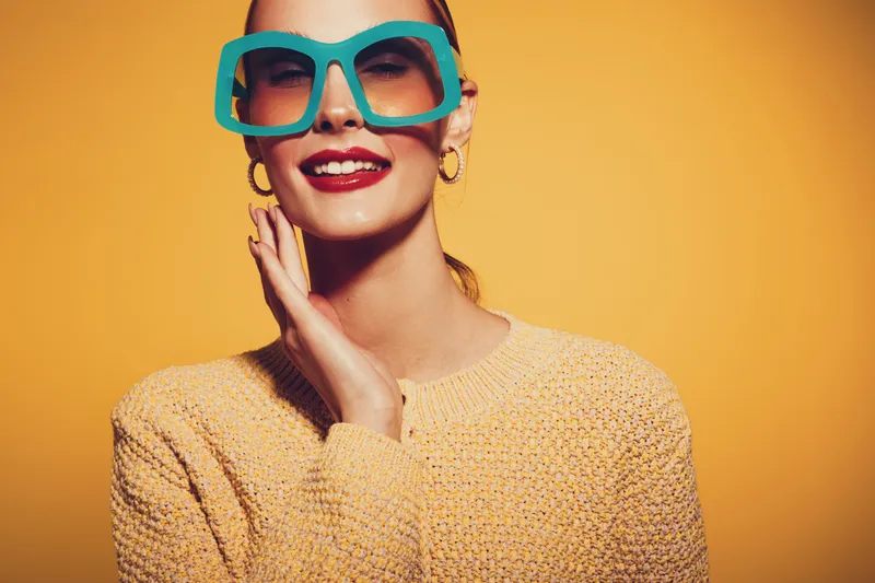 Smiling woman in oversized turquoise glasses and yellow sweater poses against orange background. Fashion portrait with bright colors and confident expression. Concept of style and joy.