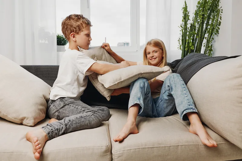 Two children playfully fight with cushions on a beige sofa in a bright living room with plants and natural light.