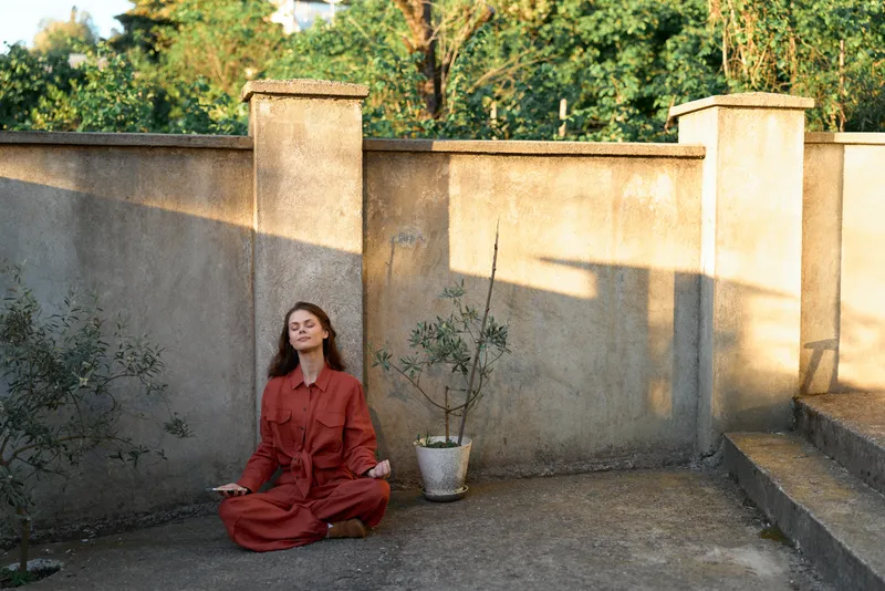 Calm woman in a red dress sits cross legged on a sunlit courtyard, embracing stillness. A minimal scene highlights self care, mindfulness, and simple daily rituals