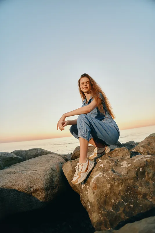 smiling woman sitting on rocks by the sea during sunset wearing casual denim overalls and sneakers with a relaxed and happy expression