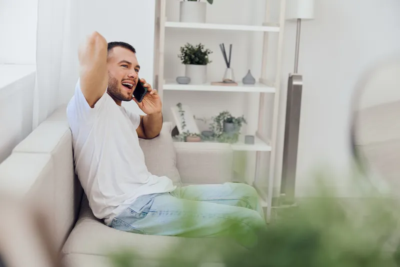 Young man happily talking on the phone while sitting on a comfortable sofa in a bright, modern living room surrounded by greenery.