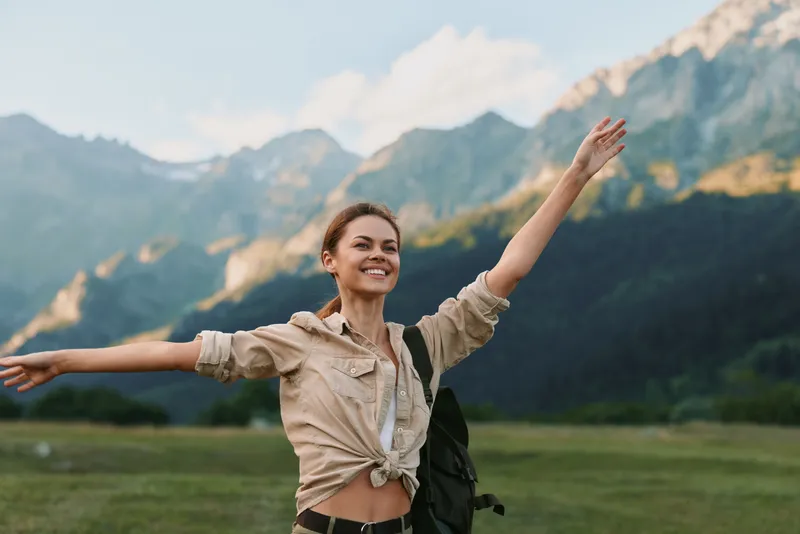 Smiling woman with outstretched arms in a scenic mountain meadow, casual beige shirt, backpack, sunlit breeze. Outdoor lifestyle, nature, wellness vibe, freedom, vitality in daylight.