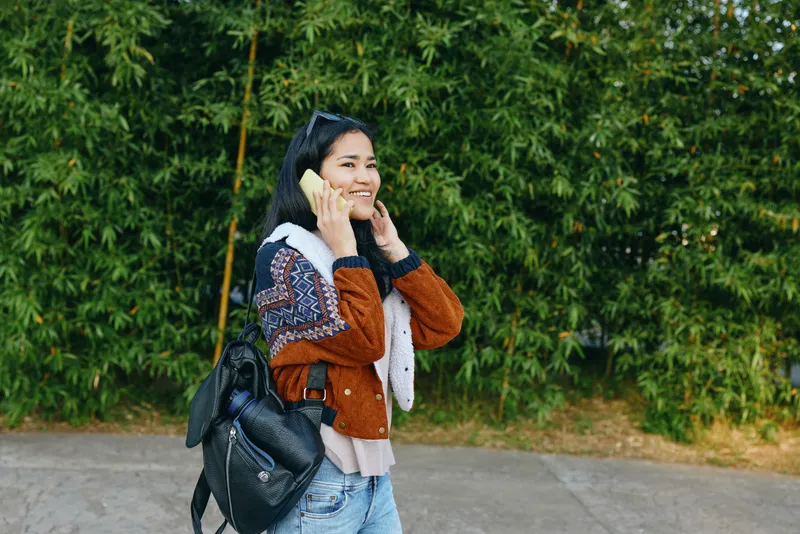 Woman talking on phone outdoors with a backpack, smiling beside a street framed by lush greenery, casual jacket and confident stance, conveying travel, communication and everyday mobility