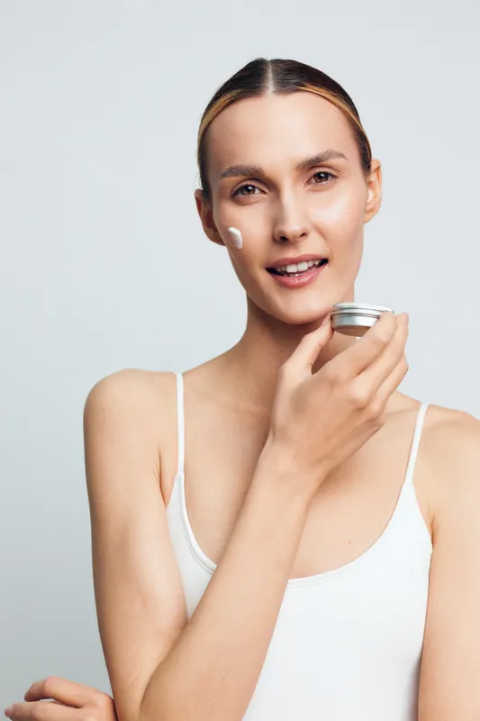 A woman applies moisturizer to her cheek in a bright studio, presenting a calm skincare routine and dewy, healthy skin with minimal makeup and soft lighting.