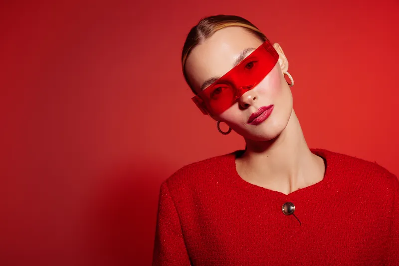 Stylish woman in red sunglasses and red outfit poses against red background. Fashion portrait with modern accessories and confident expression. Concept of bold style and elegance.