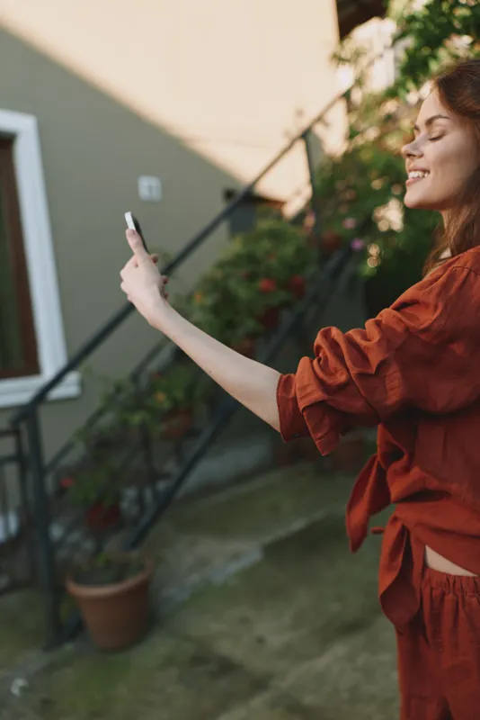 Cheerful woman outdoors, taking a selfie, smiling with sunlight on her face, casual rust outfit, relaxed pose, vibrant garden background, warm natural colors, candid lifestyle moment