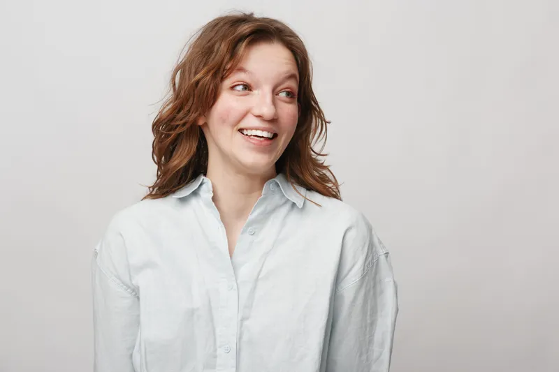 Smiling young woman with curly brown hair wearing white shirt looks to the side on light background. Concept of happiness, casual style, natural beauty, and positive emotions.