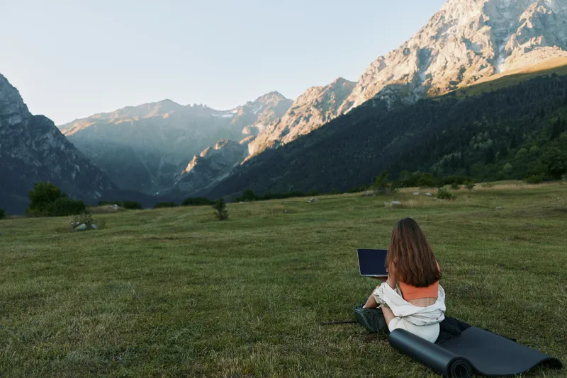 A solitary traveler sits on a portable mat in a wide meadow, working on a laptop with a dramatic mountain backdrop, blending nature and technology.