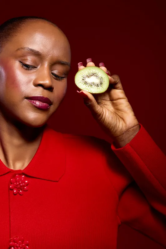 Close-up of a black African woman with dark skin holding a sliced kiwi near her face. She wears a red sweater against a red background, concept of healthy lifestyle and natural beauty.