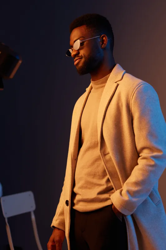 Stylish black male model in a beige coat and sweater, showcasing a trendy outfit against a colorful background, exuding confidence and modern fashion