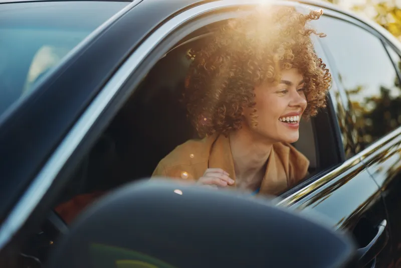 A smiling woman sits in a car, sunlight bursts through the window, curly hair glowing in warm retro film color. A candid lifestyle moment, suggesting everyday joy and freedom.