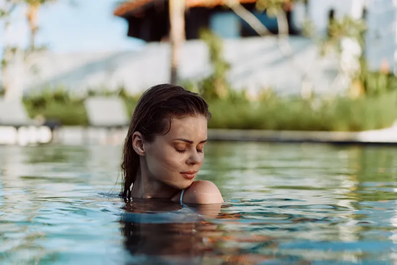 A woman relaxes in a sunlit pool, shoulders resting softly as she gazes downward. A tranquil moment of leisure and contemplation in a peaceful resort setting, capturing calm water and reflective mood.