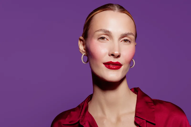 Confident woman with red lipstick and hoop earrings poses against purple background. She wears a red satin blouse and has a calm, elegant expression. Beauty and fashion concept.