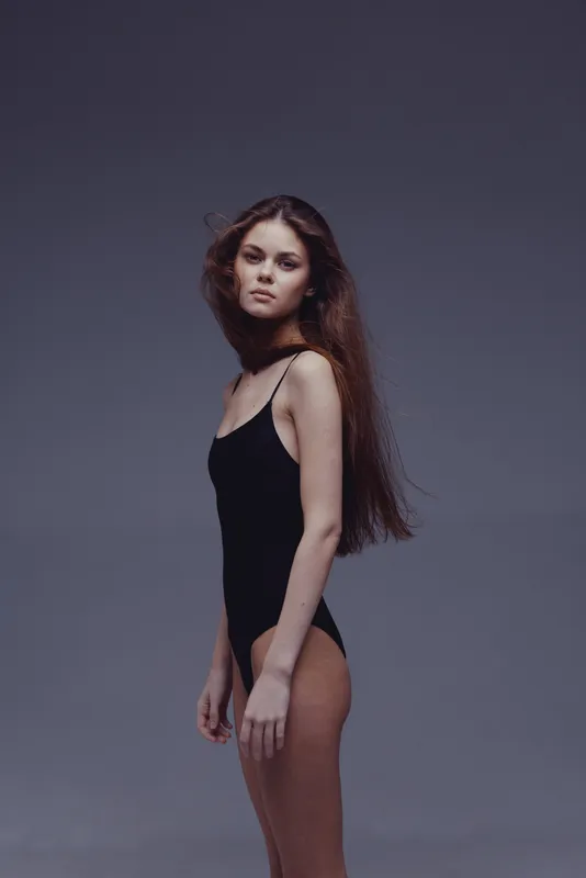 Young woman in black bodysuit posing against gray studio background with serious expression and long hair.