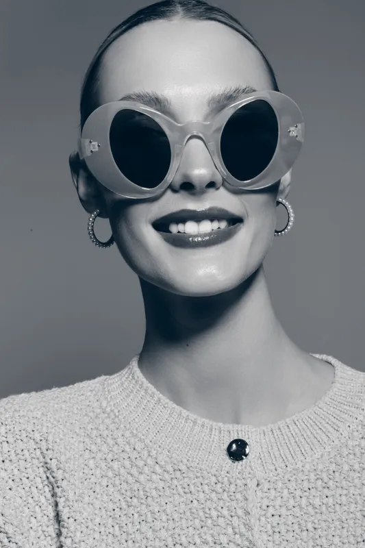 Smiling white woman with light skin wearing large round sunglasses and hoop earrings poses in knitted sweater. Concept for fashion, accessories, beauty, and lifestyle portrait.