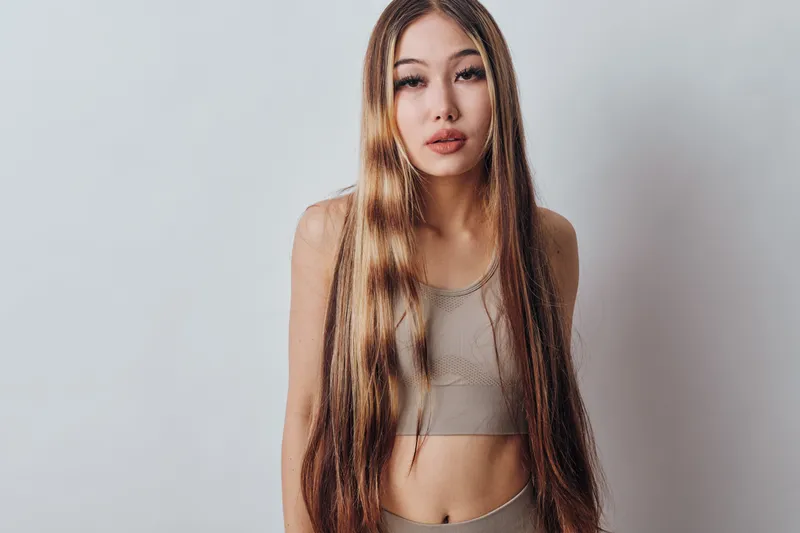 Young woman with long hair and neutral expression wearing a sleeveless top and standing against light background for fashion portrait or beauty concept.