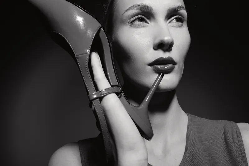 Closeup portrait of a white woman with fair skin holding red high heel shoe near face. Studio lighting highlights facial features and shoe details. Concept of fashion and beauty.