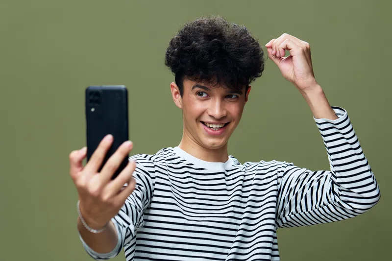 Smiling young man in striped shirt taking a selfie with smartphone, expressing happiness and excitement against green background with casual style.