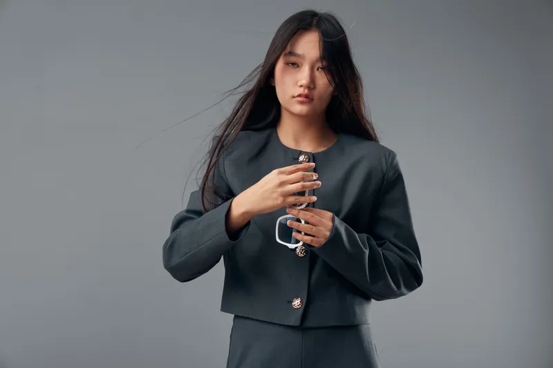 Young woman with long dark hair wearing gray jacket and pants standing against gray background with serious expression holding hands near chest with loose strands of hair.