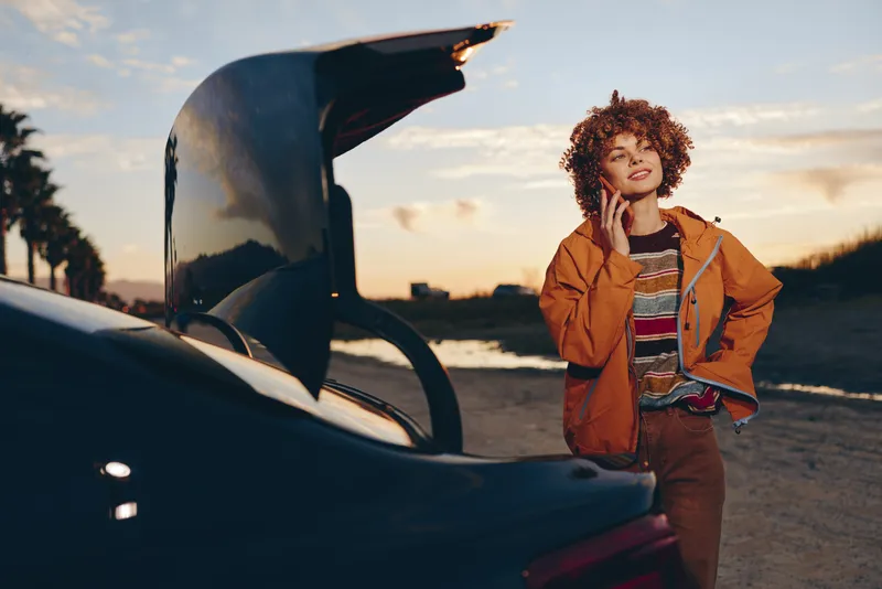 Woman lifestyle outdoors by open car at sunset, warm golden light on curly hair, orange jacket and casual jeans, relaxed stance on the road, travel mood, gentle air