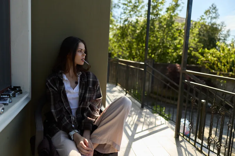 Casual woman sitting on a sunlit balcony, relaxed pose, plaid cardigan over white top, neutral tones, lush greenery in the background, a quiet lifestyle moment