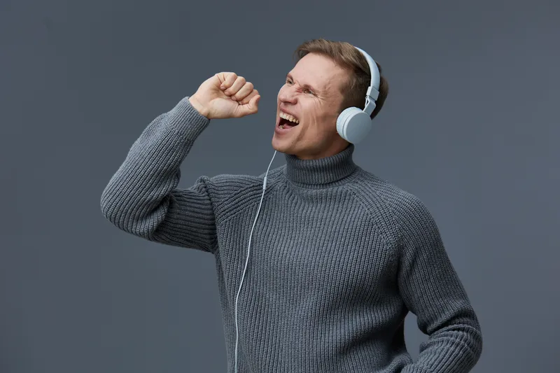 Smiling man wearing gray turtleneck sweater listening to music with headphones, enjoying music, happiness, entertainment, relaxed mood, studio background. People lifestyle concept