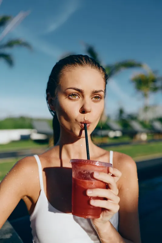 Woman enjoying an outdoor moment with a red beverage, drinking through a straw, bathed in sunlight by a poolside setting, conveying casual relaxation and summer leisure in vibrant colors