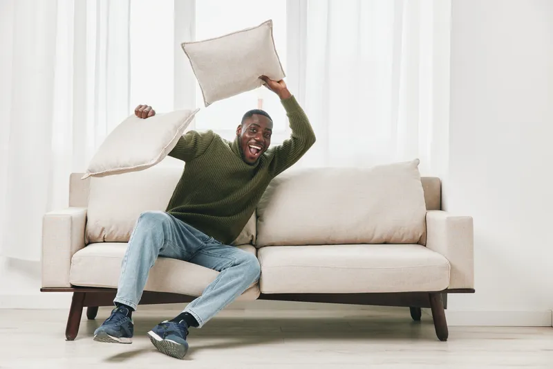 African man joyfully playing with pillows on a couch, showcasing happiness and relaxation in a bright and cozy living room setting