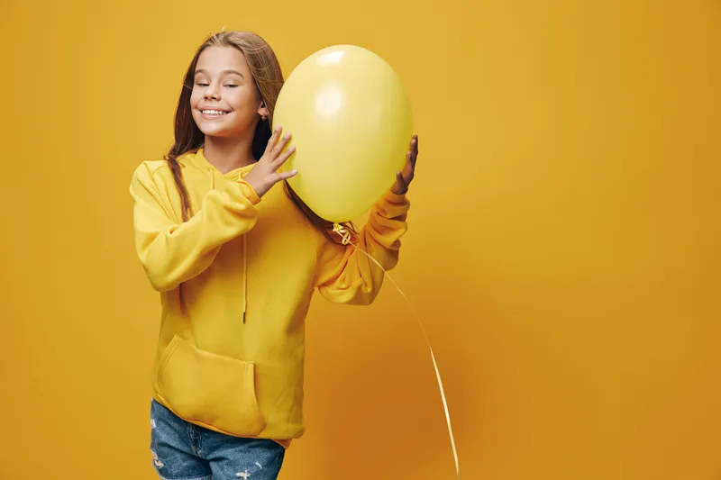 Girl smiling and holding yellow balloon against yellow background wearing casual hoodie and jeans in studio. Happy child with closed eyes and relaxed expression for celebration or joy.