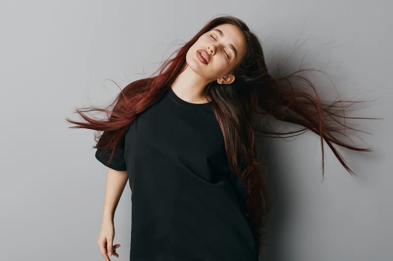 Young woman with long hair wearing black t shirt posing with eyes closed and relaxed expression against gray background in studio. Casual style and natural beauty portrait.