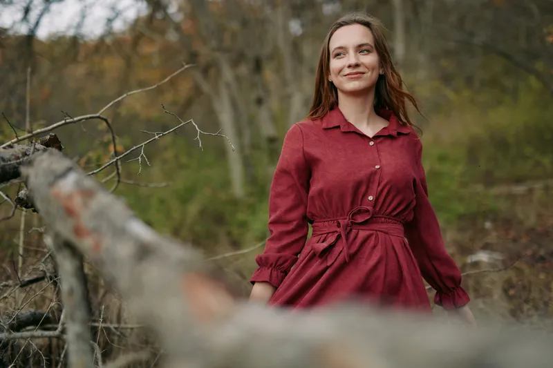 A woman in a red dress captured in an authentic, natural portrait outdoors, conveying a genuine, candid expression and calm confidence amid rustic woodland surroundings.