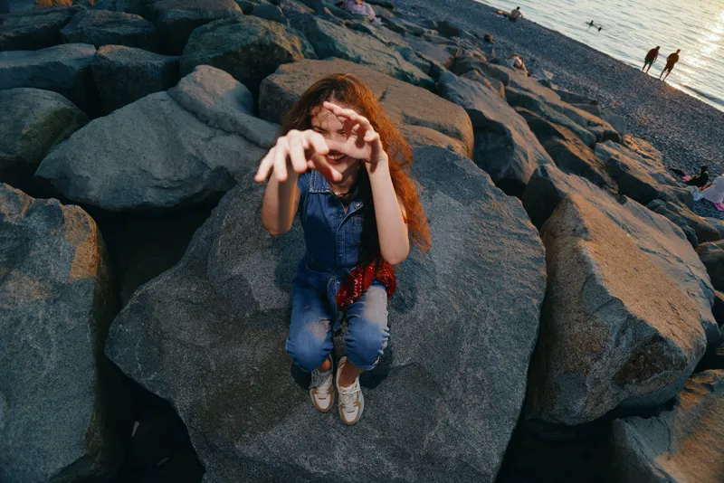 lifestyle woman in modern boho-western denim street style reaching out hands sitting on rocks by the beach in warm film color tones