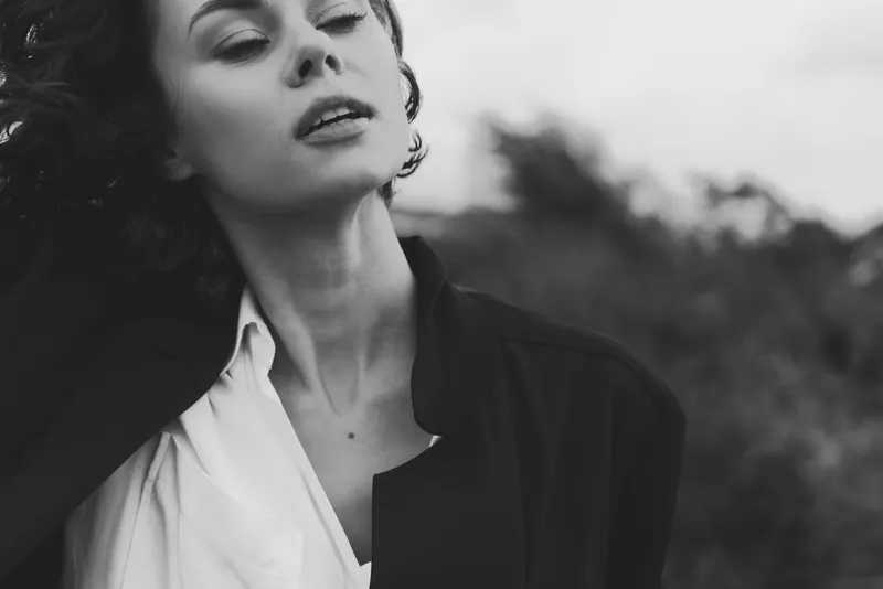 Woman in a moody outdoor scene wearing a jacket over a white shirt, wind in her hair, black and white styling, evocative fashion portrait highlighting gaze, atmosphere, and understated elegance in
