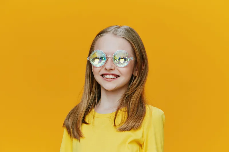 Young girl wearing stylish glasses and a bright yellow T shirt, smiling against a vibrant yellow background, embodying joy and youthful energy