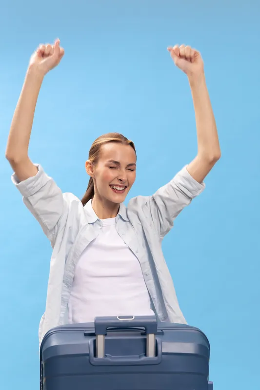 Happy young woman with suitcase raises arms in celebration, smiling with closed eyes against blue background. Concept of travel, vacation, freedom, and joyful moments.