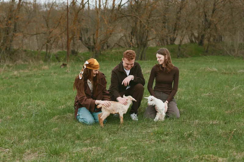 Three friends outdoors in a green field interact with two young goats, smiling and petting them. A warm, candid lifestyle moment amid countryside scenery and natural light, capturing companionship