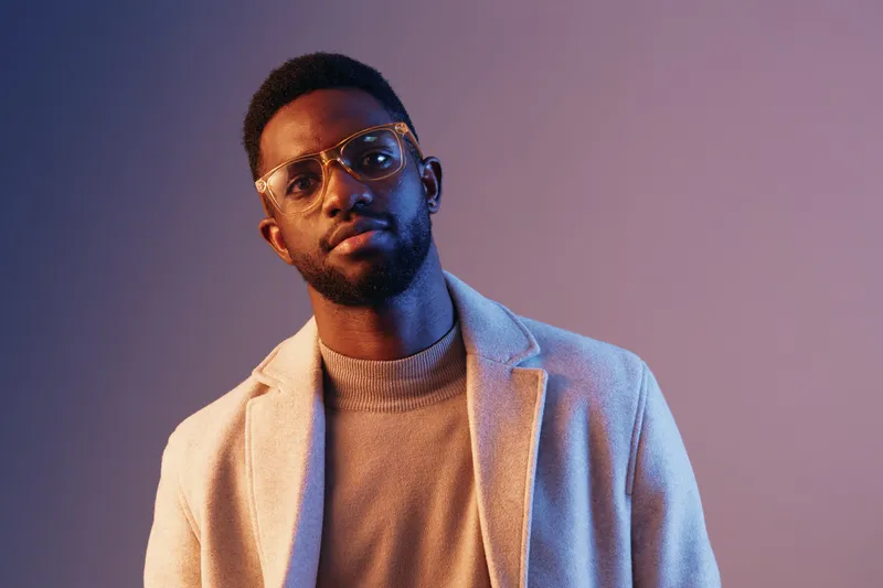 Stylish young African man in glasses wearing a modern sweater and coat, posing confidently against a soft purple background, reflecting a trendy fashion concept