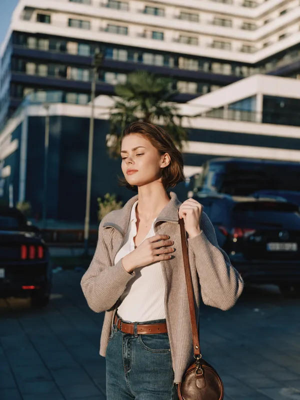 A stylish urban woman adjusts her coat on a sunny street, carrying a bag as she strides past modern buildings, exuding confidence and contemporary fashion.