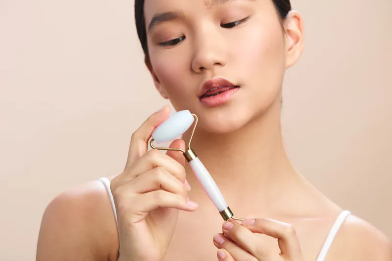 Asian woman gently uses a facial roller on her cheek in a softly lit studio, presenting a calm skincare moment that emphasizes self care, balance, and mindful beauty.
