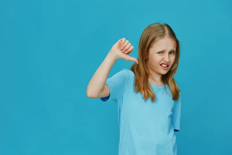 Young girl showing a thumbs down gesture against a bright blue background, expressing disappointment with a playful facial expression while wearing a casual blue shirt