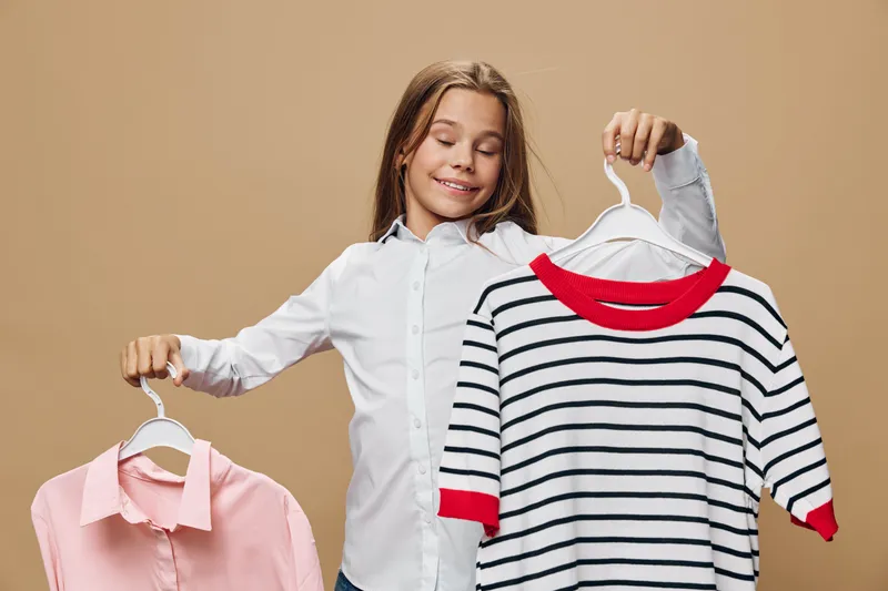 Girl holding two clothes hangers with casual tops smiling in studio, choosing outfit, comparing fashion styles, clothing, youth, lifestyle and wardrobe selection concept.