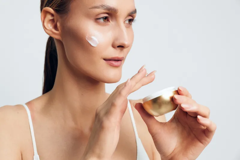 Young woman with clear skin applying white face cream on cheek. Close-up portrait with natural light. Concept of skincare, beauty routine, and healthy lifestyle.