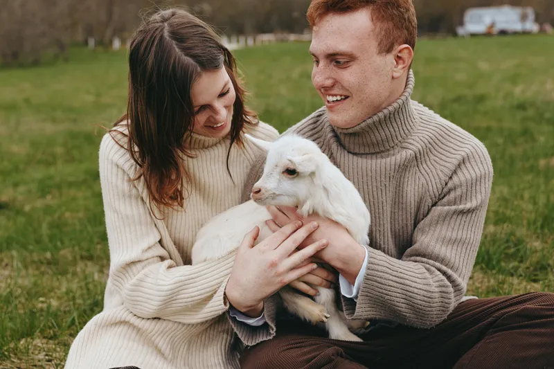 Happy couple in cozy sweaters cuddle a young goat kid outdoors, sharing smiles in a countryside field, creating a warm lifestyle moment, gentle light, affectionate atmosphere, peaceful companionship,