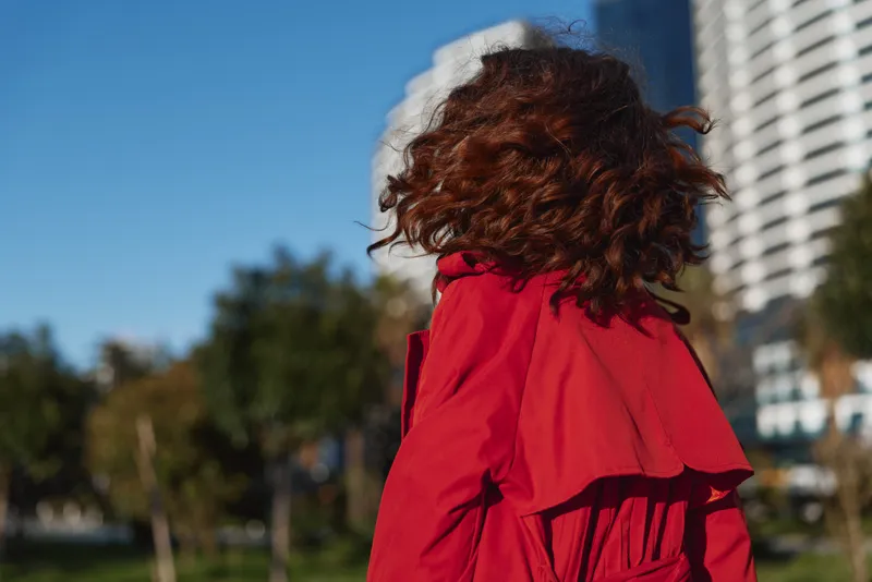 Young woman with curly hair wearing a red coat strolls through a sunny city park with modern high rise buildings in the background, showcasing street style.