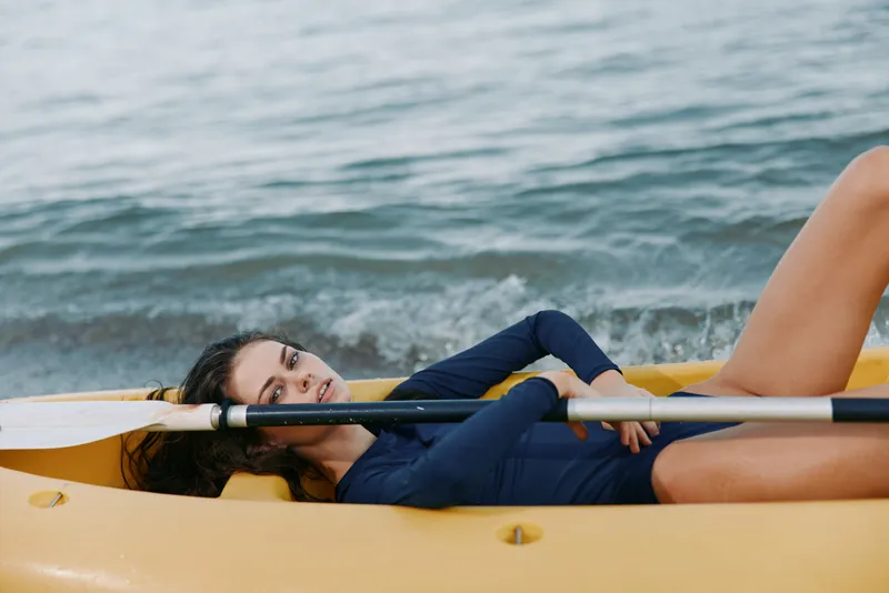A woman reclines inside a yellow kayak on calm water, wearing a navy swimsuit, resting a paddle across her chest and gazing toward the camera with a relaxed, serene expression.
