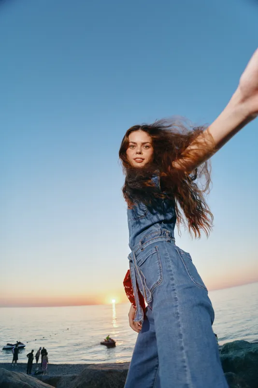 Lifestyle woman in modern boho-western denim street style poses confidently by the ocean at sunset with film color tones, capturing relaxed and natural vibes.